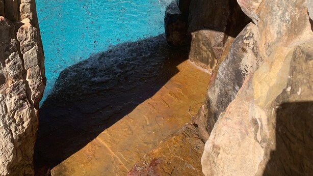 A stone-walled pool with clear blue water, surrounded by rocks and a view of outdoor seating.