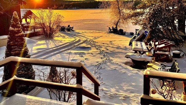 A snowy outdoor area with a wooden deck, a snow-covered Christmas tree, and a view of a lake during sunset. -  Newly remodeled-Lakefront cabin on 4th Lake w/dock & kayaks. Trail 5 Access (Old Forge)