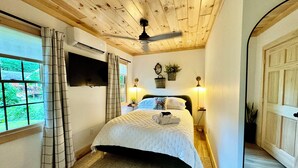 A bedroom with a wooden ceiling, a bed with a white quilt, a TV mounted on the wall, and a window with curtains. -  Newly remodeled-Lakefront cabin on 4th Lake w/dock & kayaks. Trail 5 Access (Old Forge)