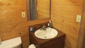 Une salle de bain aux murs lambrissés, avec un lavabo blanc, un miroir et des articles de toilette posés sur le comptoir.