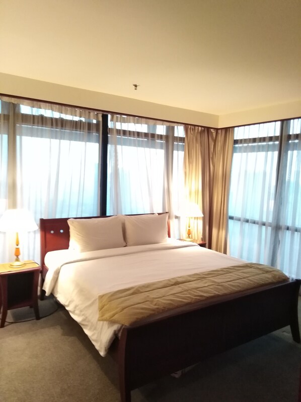 A hotel room with a large bed, two bedside tables with lamps, and a view through sheer curtains.
