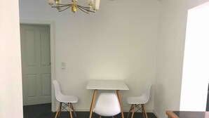 Un coin repas avec une table blanche et quatre chaises blanches aux pieds en bois, un lustre et un poêle.