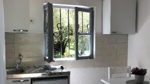 A compact kitchen with a built-in fridge, a window with a view of greenery, a dining table with chairs, and a hanging light fixture. - Rajana bungalow 2(Olive House) (Ulcinj)