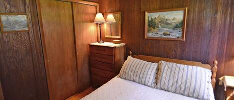 A bedroom with wooden paneling, a bed with striped bedding, a nightstand with lamps, and a painting on the wall.