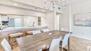 Un comedor moderno con una gran mesa de madera, sillas blancas, una araña y una cocina al fondo.