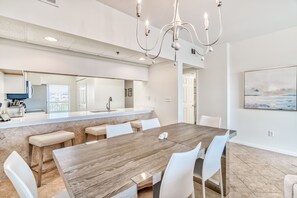 Ein moderner Essbereich mit einem großen Holztisch, weißen Stühlen, einer Chandelierleuchtre und einer Küche im Hintergrund.