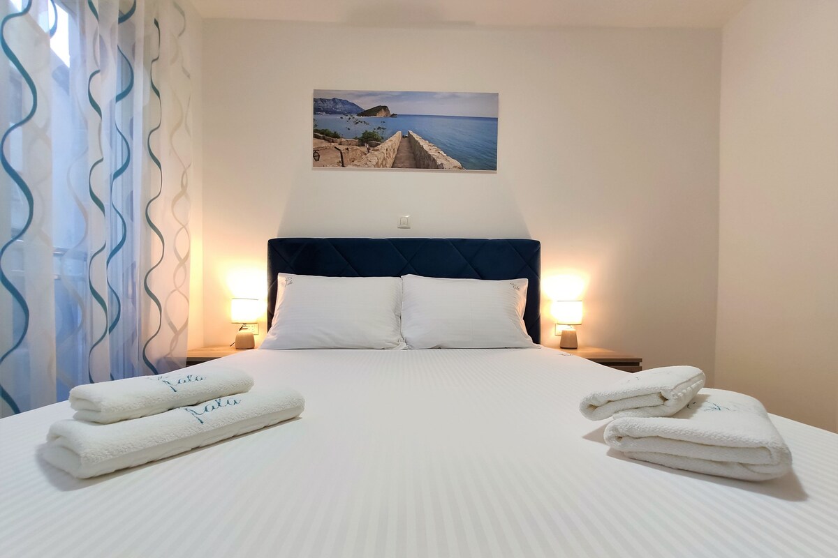 Une chambre d’hôtel avec un lit, deux lampes de chevet, un tableau représentant un paysage côtier et deux serviettes sur le lit.