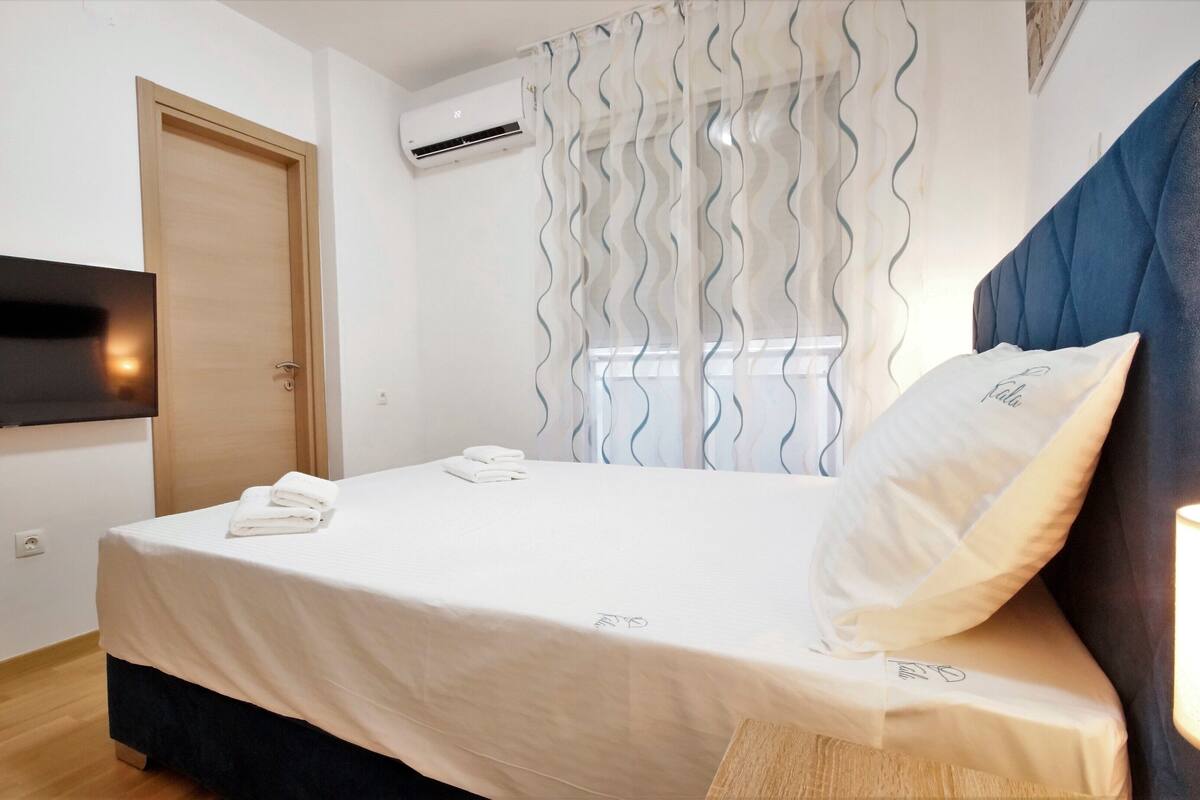 Una habitación de hotel con cama, televisor montado en la pared, aire acondicionado y ventana con cortinas.