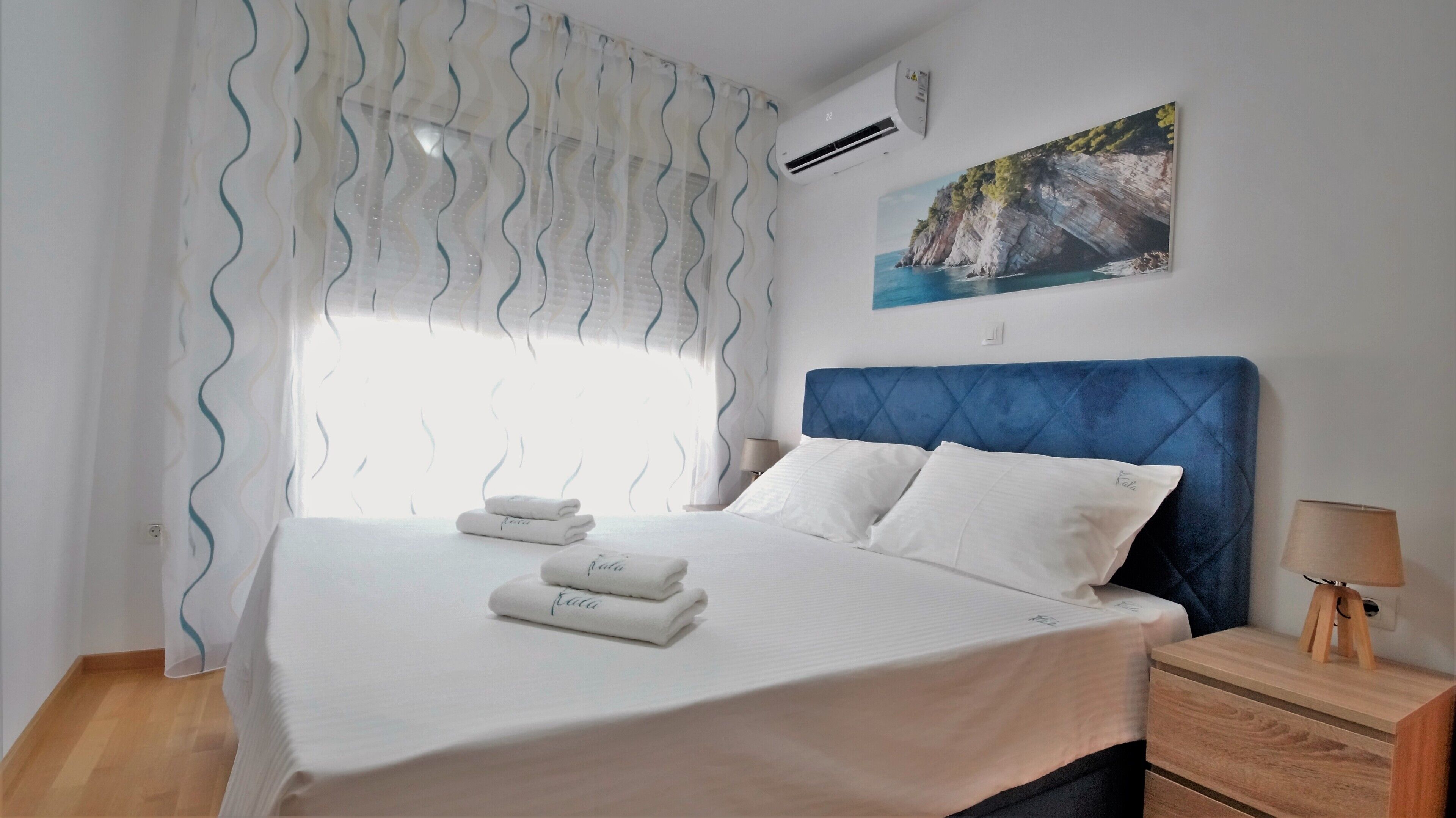 A hotel room with a bed, a nightstand, a lamp, a painting, and an air conditioning unit.