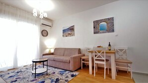 Una sala de estar con un sofá color beige, una mesita redonda, una mesa de comedor con sillas, un reloj de pared y dos cuadros.