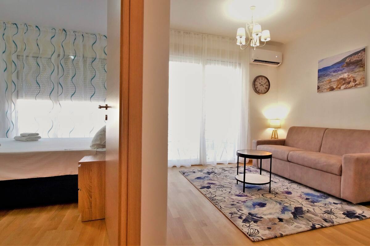 Un dormitorio con cama, mesita de noche, ventana con cortinas y vistas al exterior.