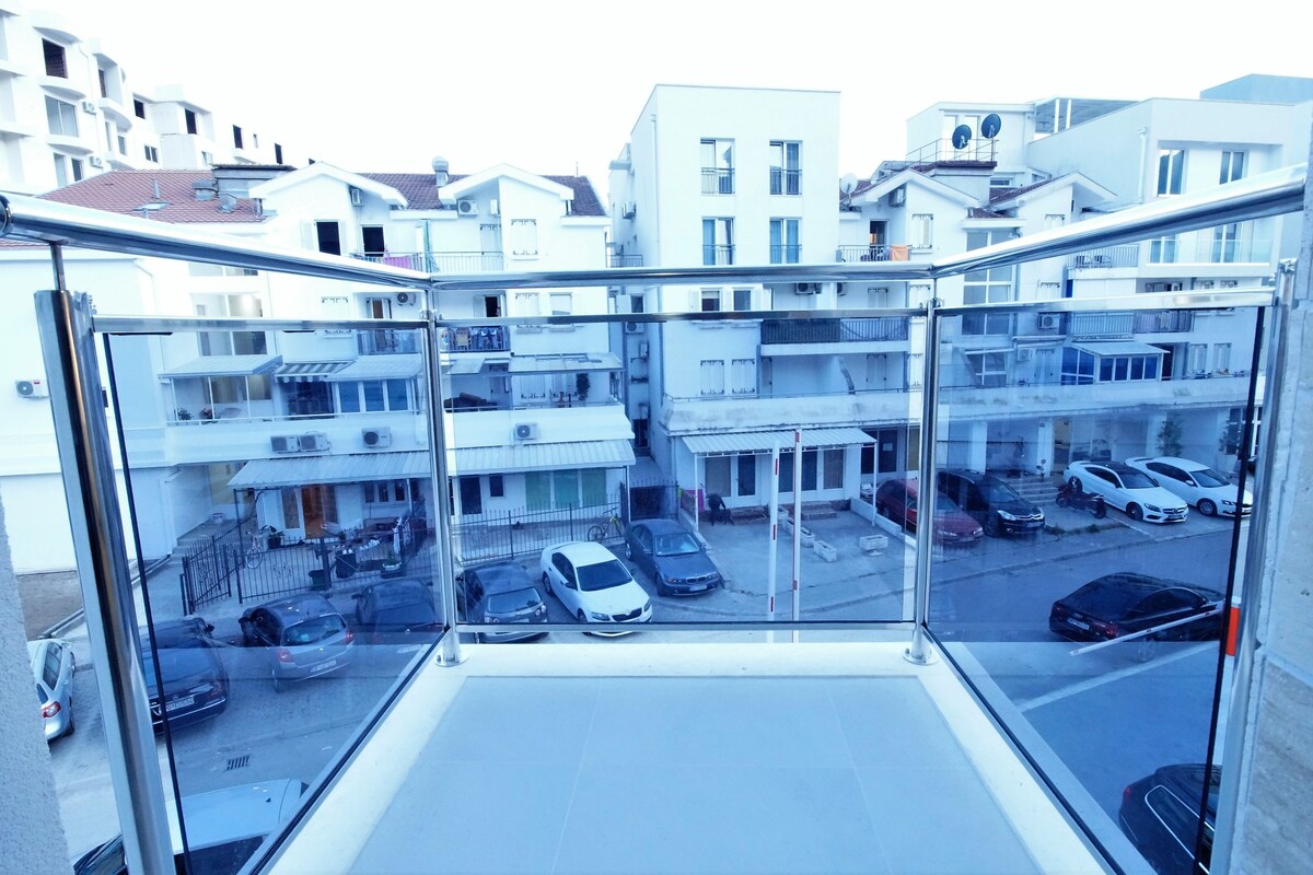 Un appartement situé dans un immeuble de grande hauteur, doté d’un balcon donnant sur un parking et les bâtiments avoisinants.