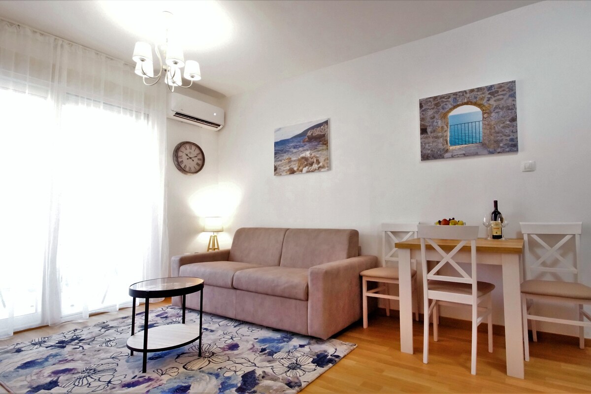 Un salon avec un canapé beige, une petite table ronde, une table à manger avec des chaises, une horloge murale et deux tableaux.