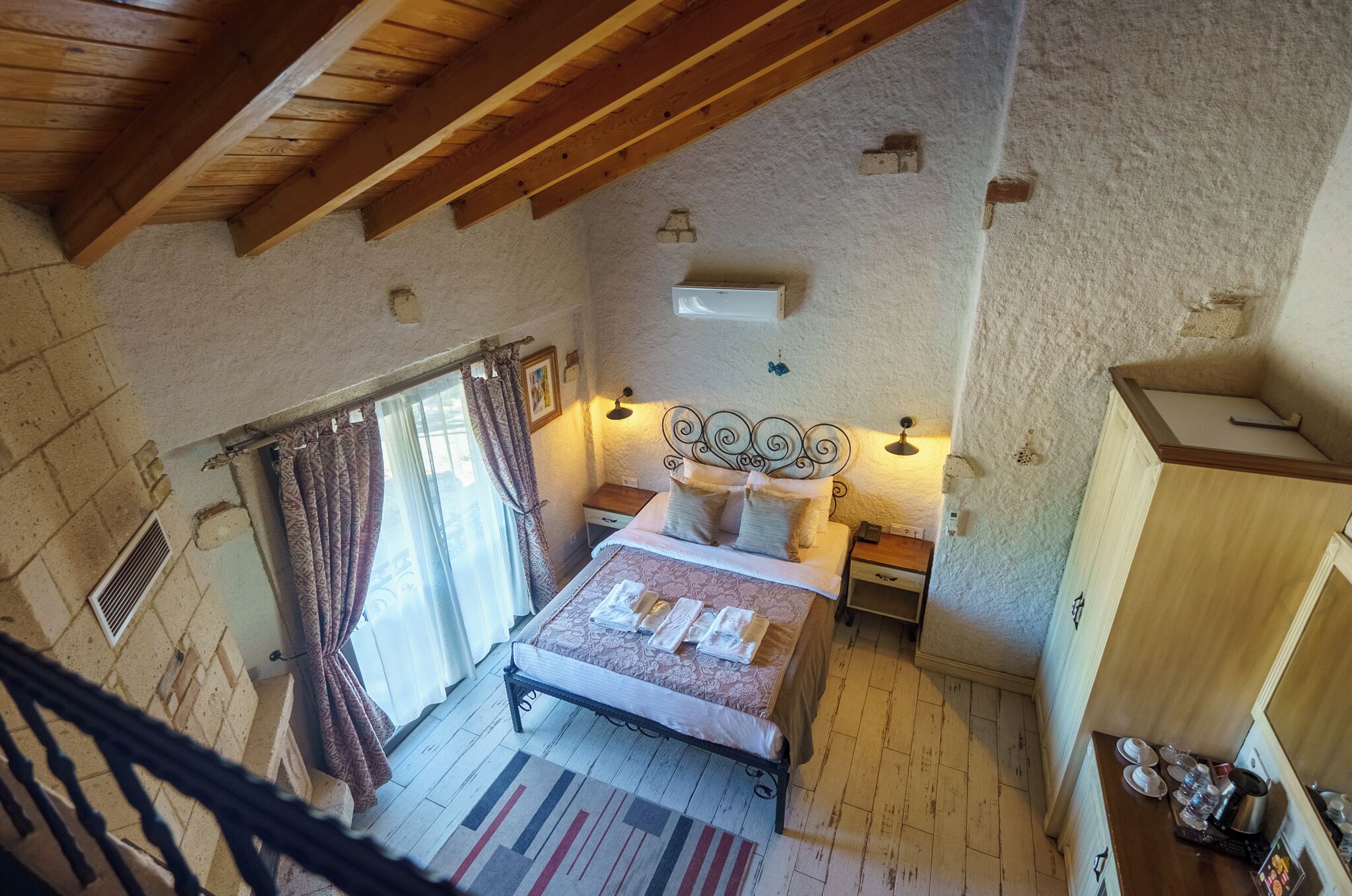 A bedroom with a wooden ceiling, a bed with a patterned bedspread, a nightstand with a lamp, and a window with curtains.