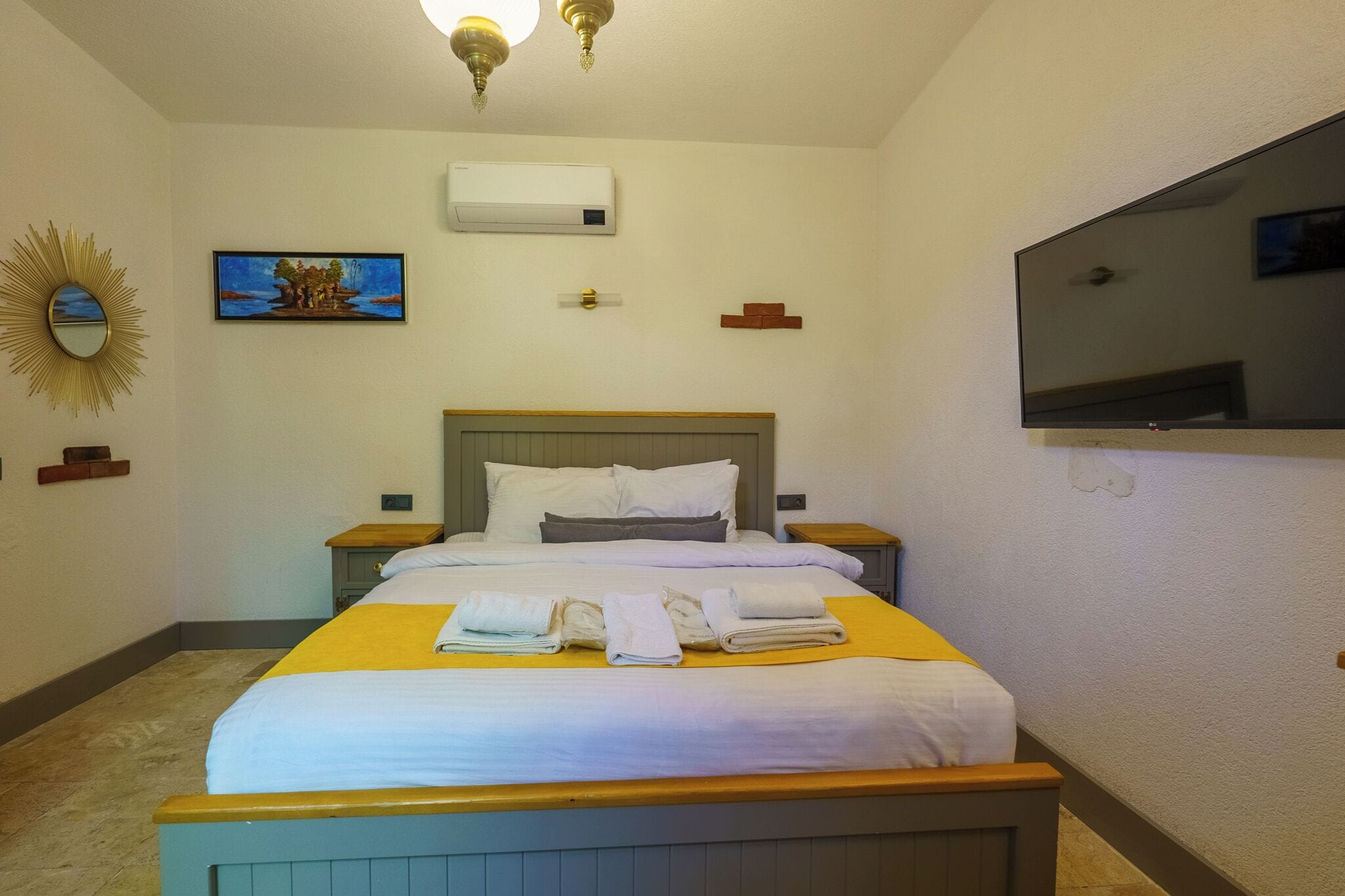 A hotel room with a bed, bedside tables, a flat-screen TV, an air conditioning unit, a painting, and a mirror.