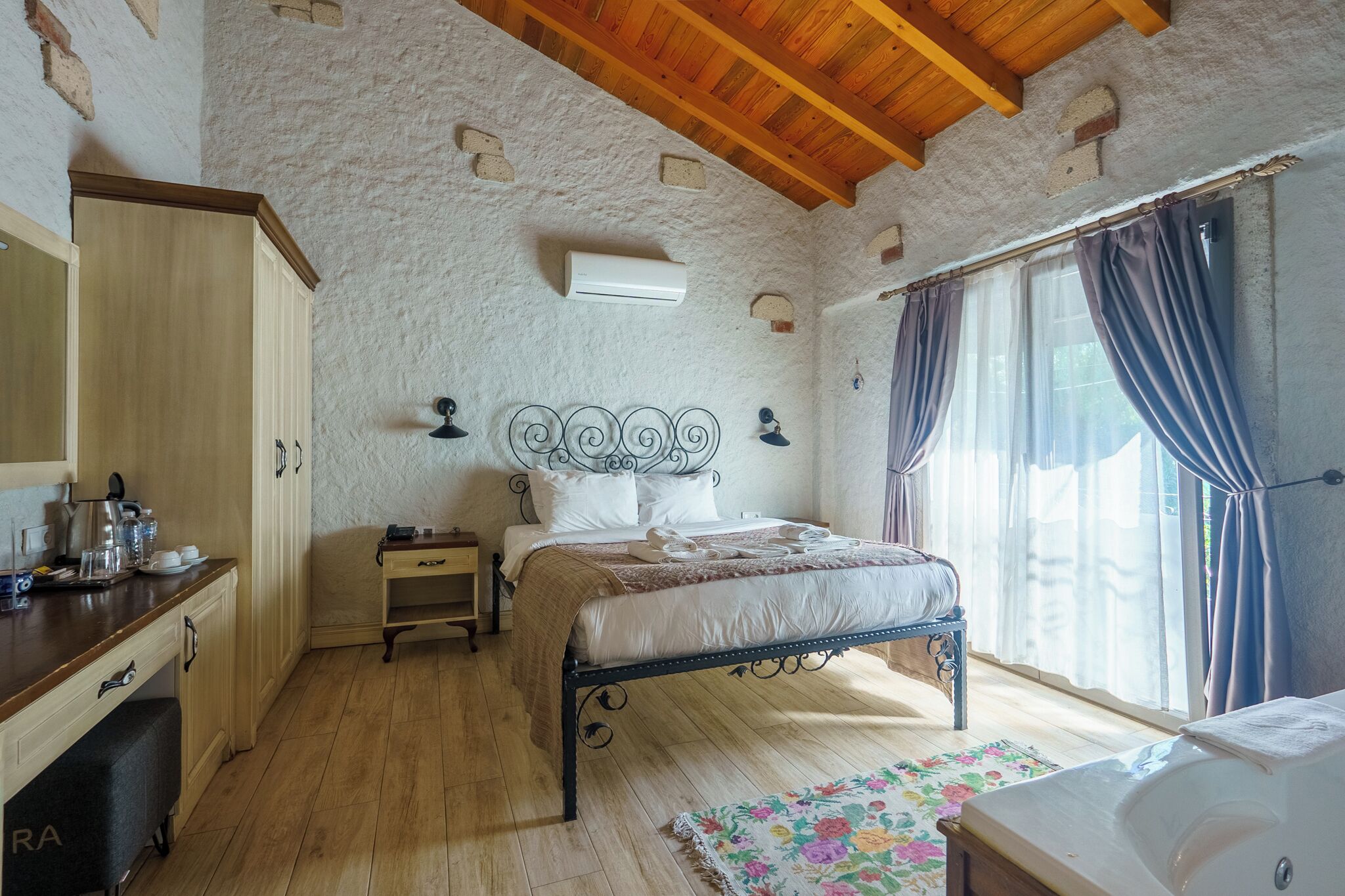 A bedroom with a wooden ceiling, a bed with a metal frame, a bedside table, a nightstand, and a window with curtains.