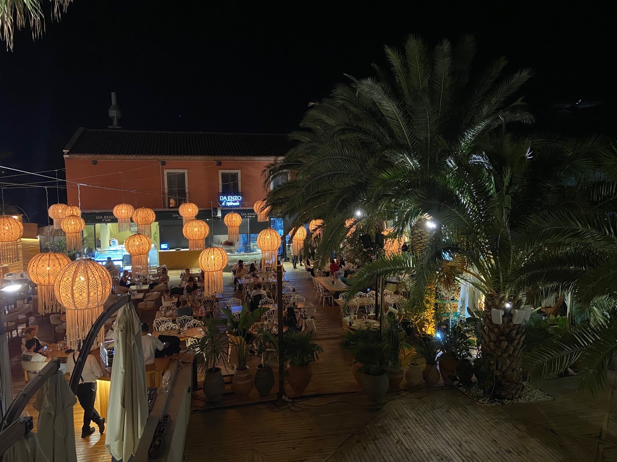 An outdoor dining area with tables and chairs, palm trees, and hanging lanterns.