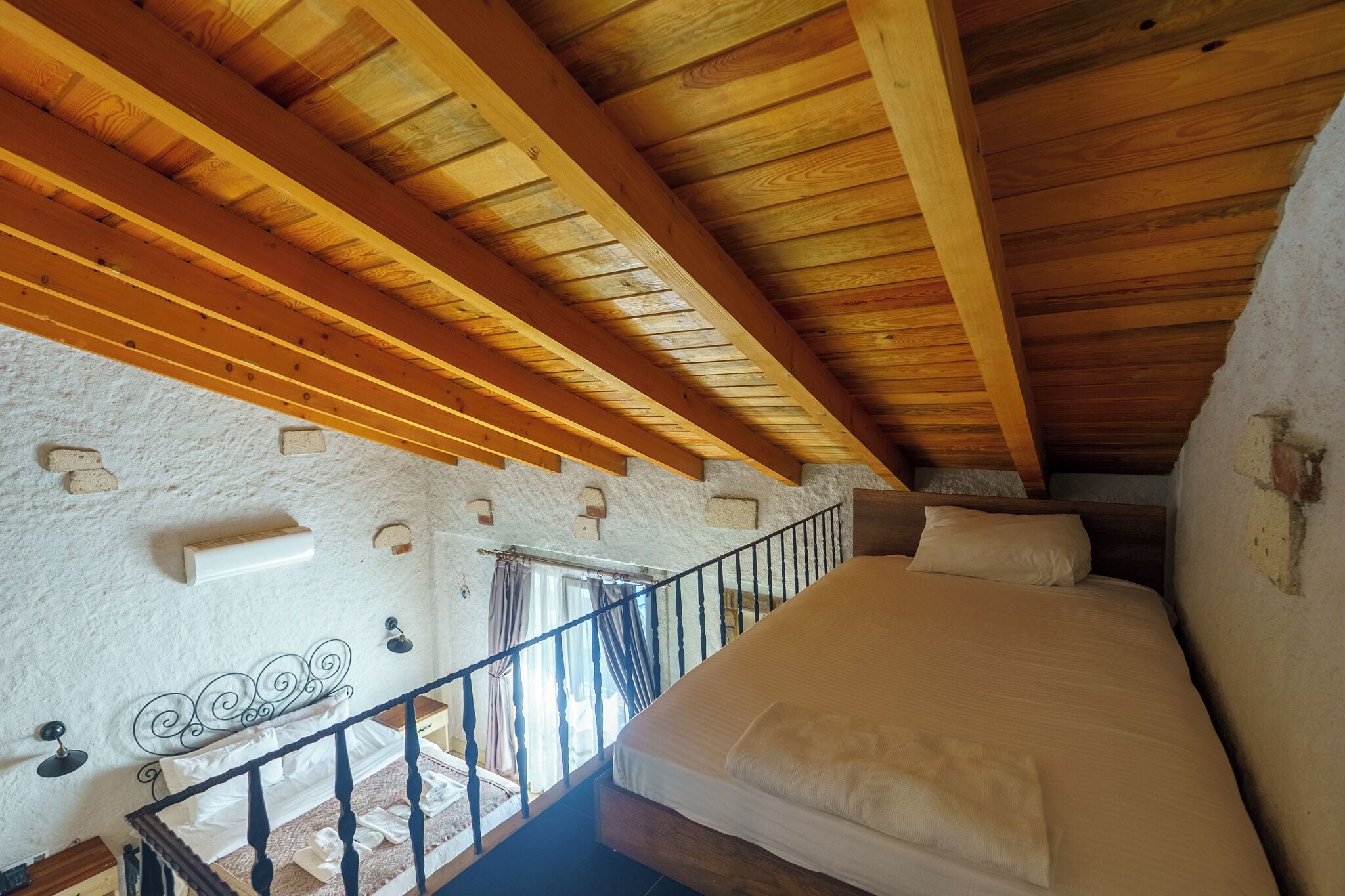 Ein Schlafzimmer mit Holzbalkendecke, einem Bett, einem Fenster mit Vorhängen und einer weiß strukturierten Wand.