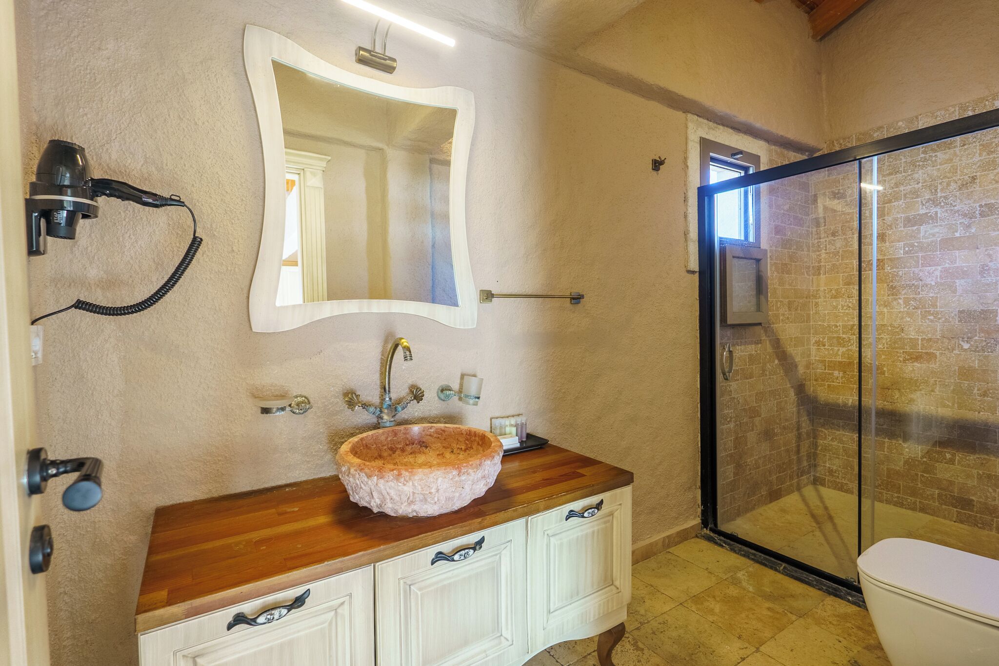 A bathroom with a wooden vanity, a round sink, a mirror, a hairdryer, a glass shower enclosure, and a toilet.