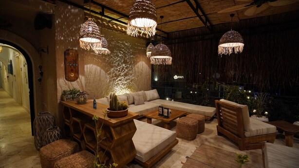 A cozy lounge area with a wooden sofa, cushioned chairs, and a wooden coffee table. There are hanging pendant lights and a large window with a view of the city. - Alacati Aura Plus (Cesme)