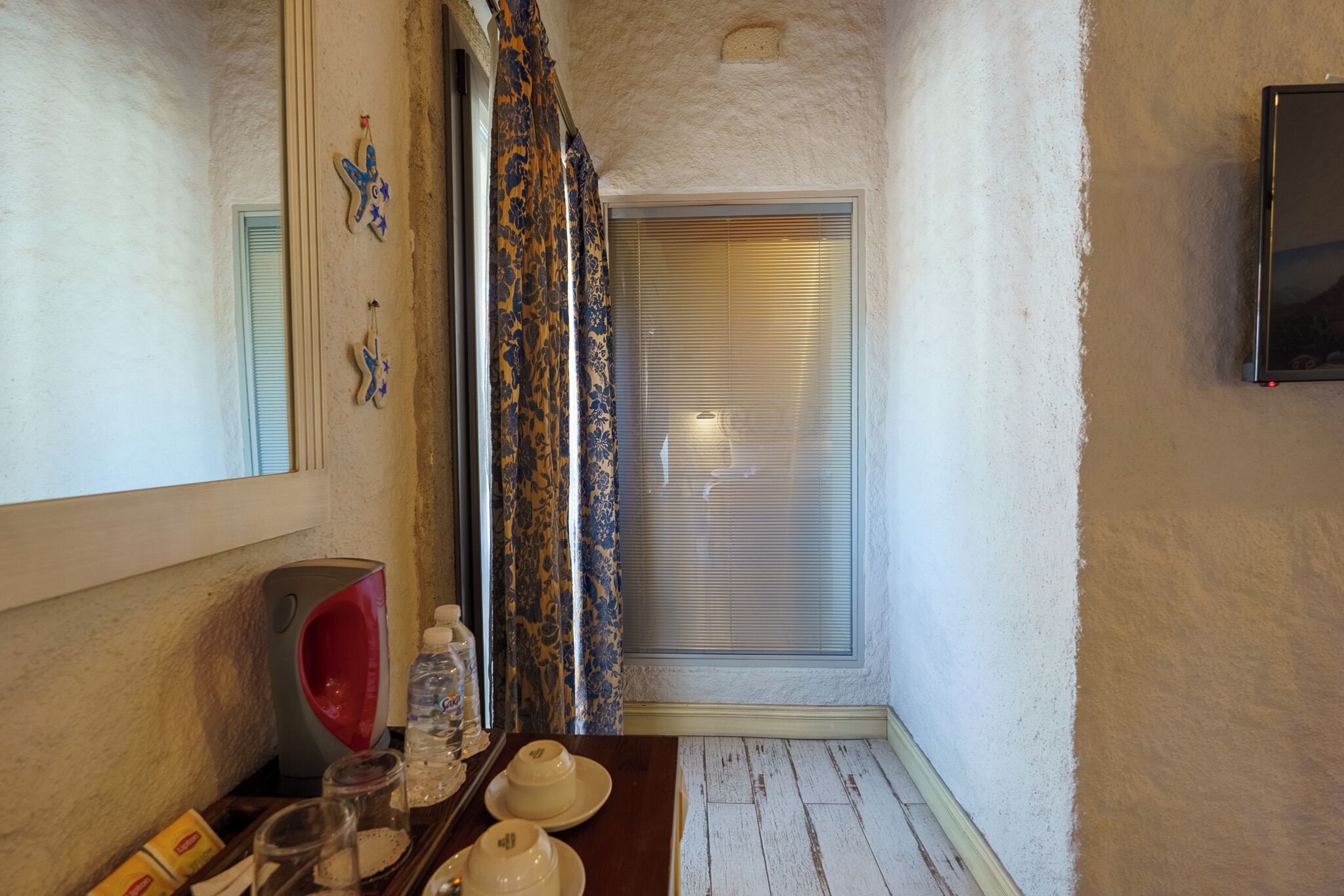 Una esquina de una habitación con ventana, un espejo, una mesa con una tetera, una botella de agua y un salero.