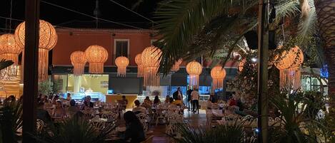 Un espace de restauration en plein air, avec des tables et des chaises, entouré de palmiers et agrémenté de guirlandes lumineuses décoratives.