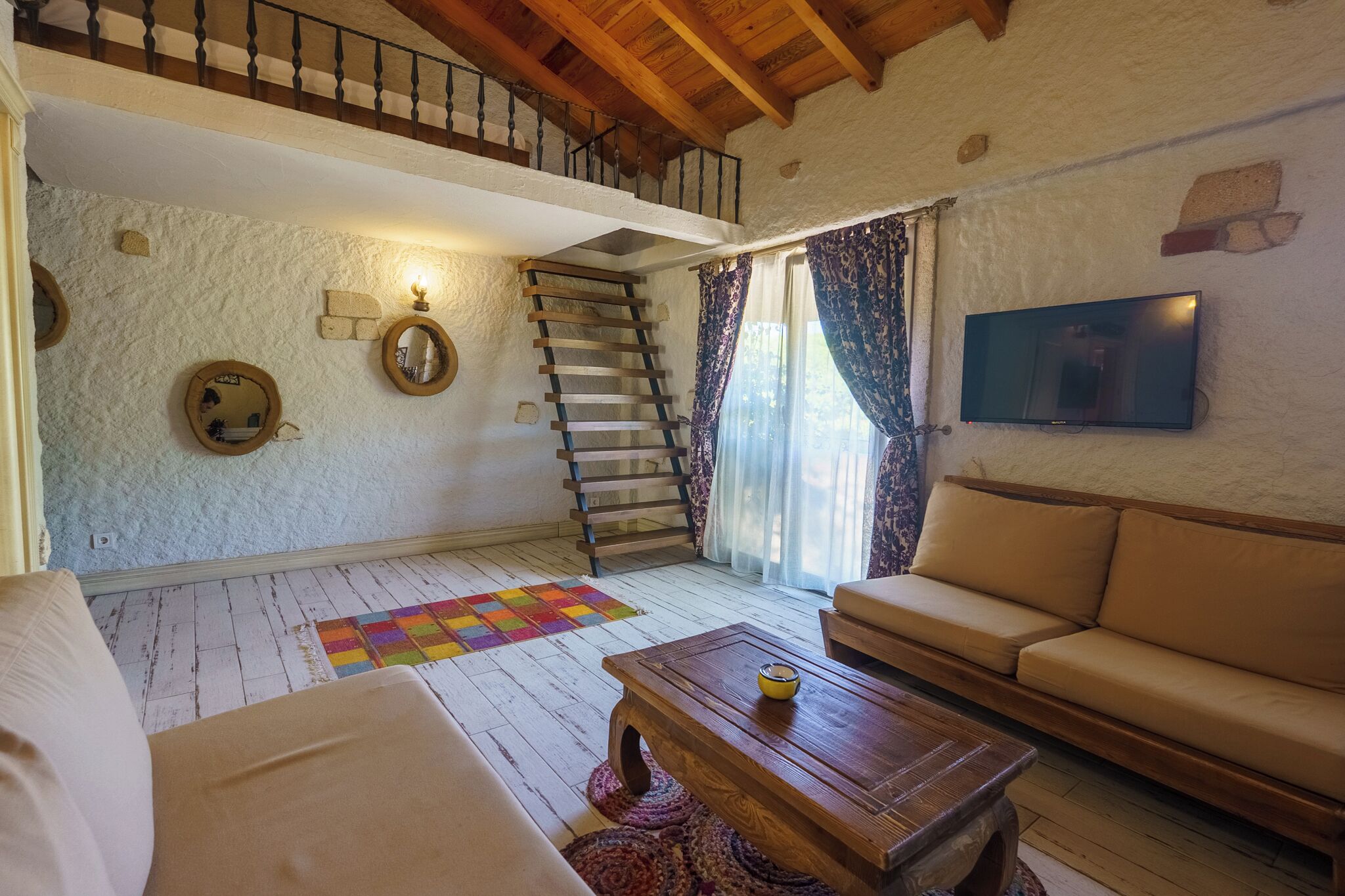 Ein gemütliches Wohnzimmer mit einer Holztreppe, einem Sofa, einem Couchtisch und einem an der Wand montierten Fernseher.