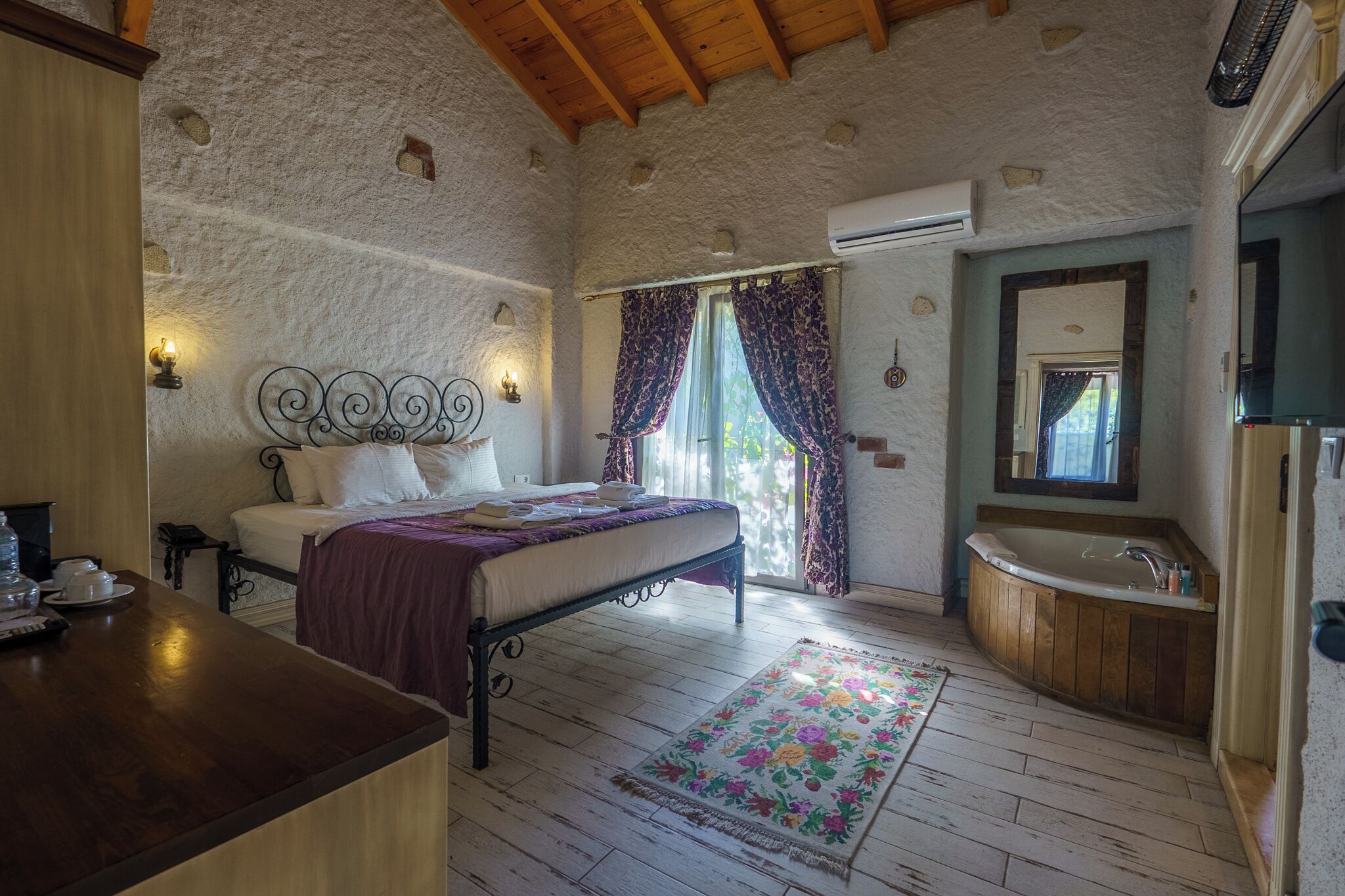 A bedroom with a bed, a wooden desk, a bathroom with a bathtub, and a large window with curtains.