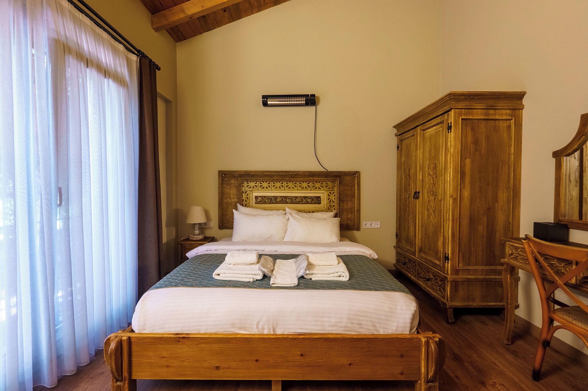 A neatly made bed with white linens and towels, a wooden headboard, a wooden wardrobe, and a desk with a chair.