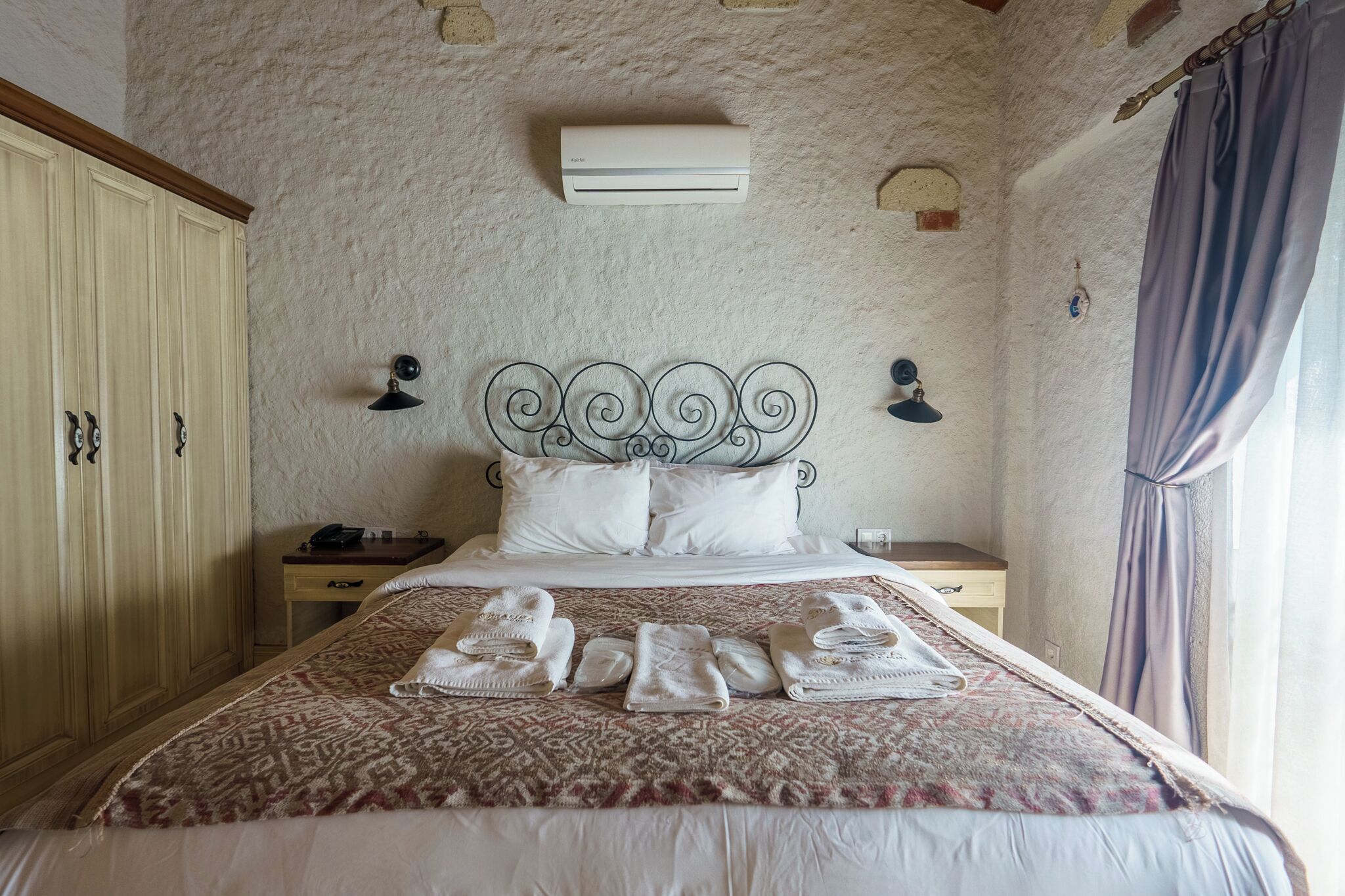 Un lit bien fait, recouvert d’une couvre-lit à motifs et agrémenté de plusieurs serviettes, une table de chevet avec une lampe, une armoire et un climatiseur fixé au mur.