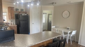 Cocina con encimera de granito, refrigerador negro y armarios de madera. Hay un comedor con una mesa y sillas blancas.