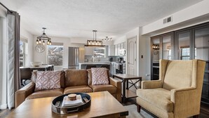 A modern living room with a leather sofa, a wooden coffee table, and a beige armchair. A kitchen with white cabinets and stainless steel appliances is visible in the background.