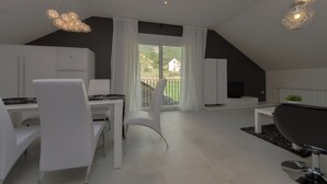Een moderne, goed verlichte eetruimte met witte meubels, een groot raam met gordijnen en uitzicht op groen en een huis.