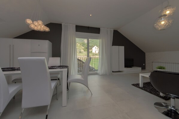A modern, well-lit dining area with white furniture, a large window with curtains, and a view of greenery and a house outside.