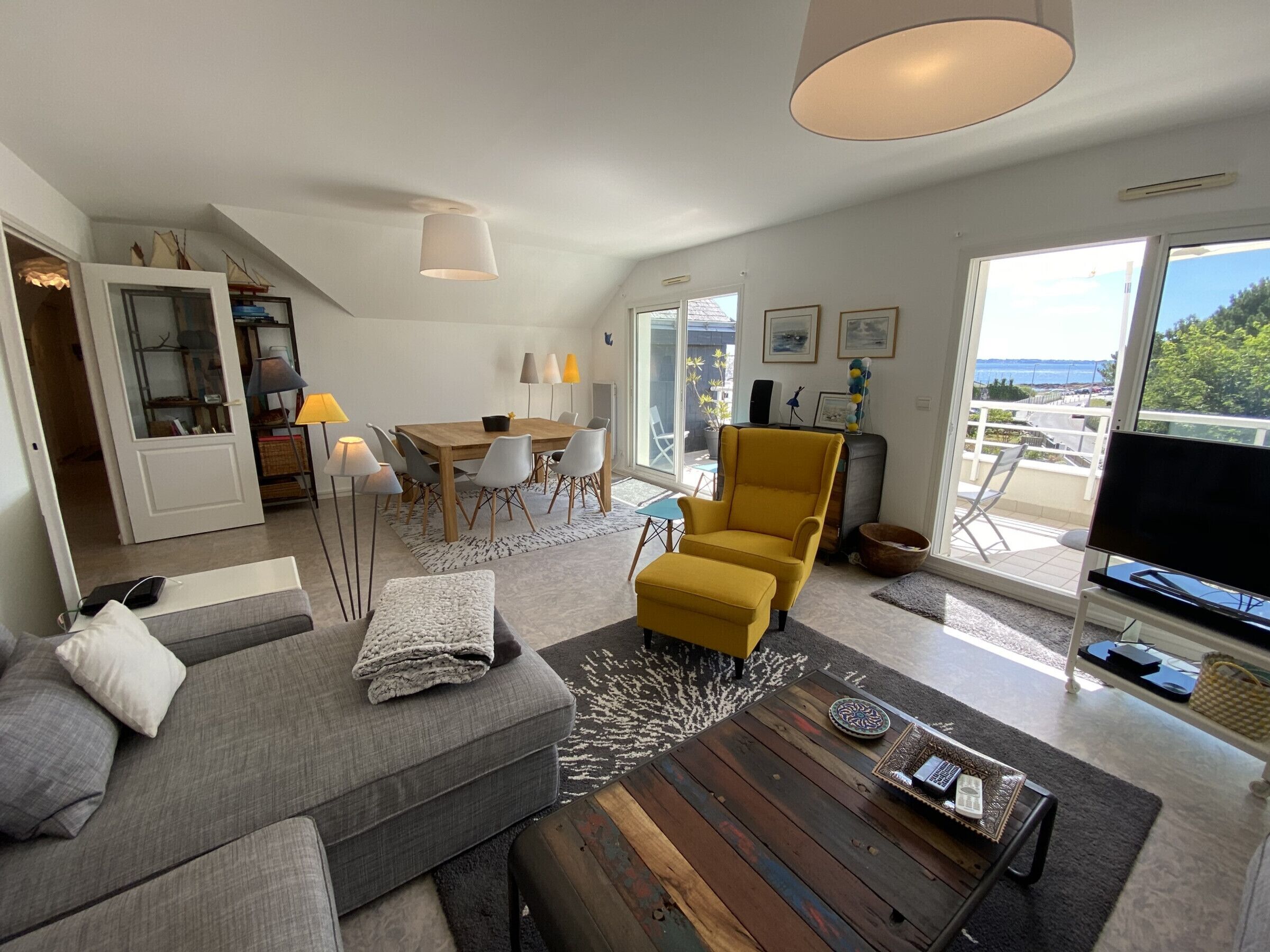 A modern living room with a sofa, ottoman, coffee table, and a yellow armchair. There is a dining area with a table and chairs, and a balcony with a TV.