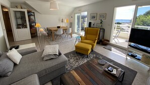 A modern living room with a sofa, ottoman, coffee table, and a yellow armchair. There is a dining area with a table and chairs, and a balcony with a TV.