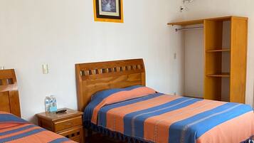 Un dormitorio con dos camas, un armario de madera, una mesita de noche pequeña, un ventilador de pared y un cuadro enmarcado.