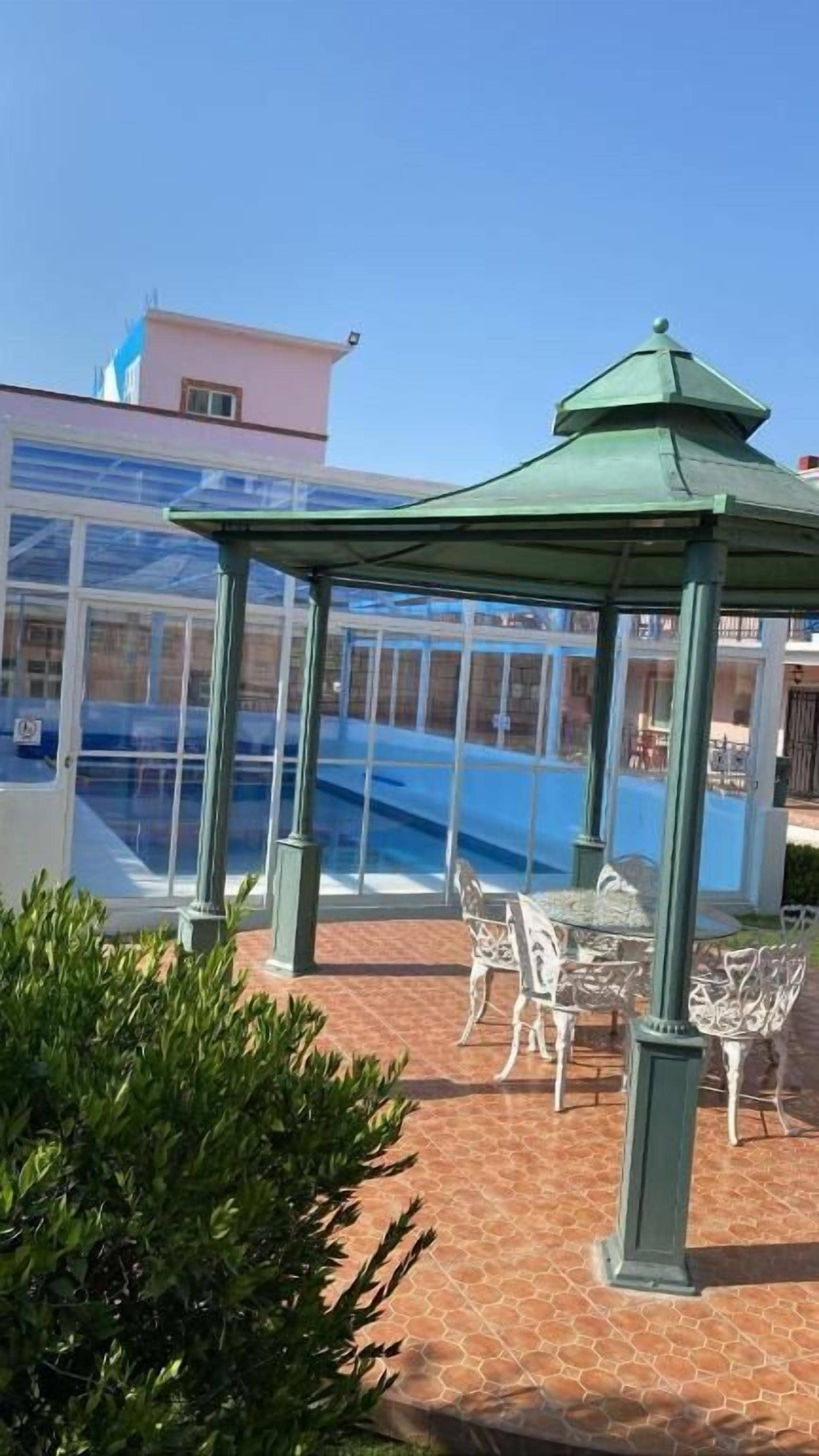 A glass-enclosed gazebo with a green roof, a pool, and outdoor furniture.
