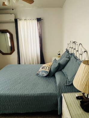 A bedroom with a bed, a bedside table, a lamp, a mirror, and a window with curtains. - Cozy Caribbean Home Near Beaches (Christiansted)