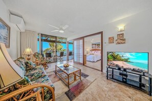 A living room with a sofa, chairs, a coffee table, a TV, and a sliding glass door leading to a balcony with a view of the sea.