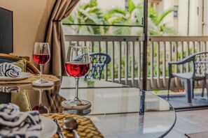 Una mesa de vidrio con dos copas de vino y platos, una cesta de mimbre y vistas a un balcón con sillas y follaje.