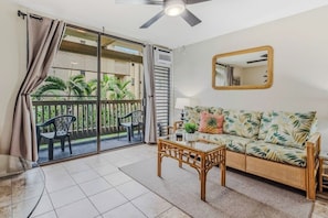 A room with a sliding glass door leading to a balcony, a wicker sofa, a small table, and a ceiling fan. - Delightful condo near beaches with pool & hot tub & private balcony (Kailua-Kona)