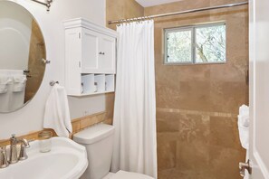 A bathroom with a white sink, a toilet, a shower with a glass door, and a window with a view of trees. - Blue Spring Retreat I Hot tub, Families (Wimberley)