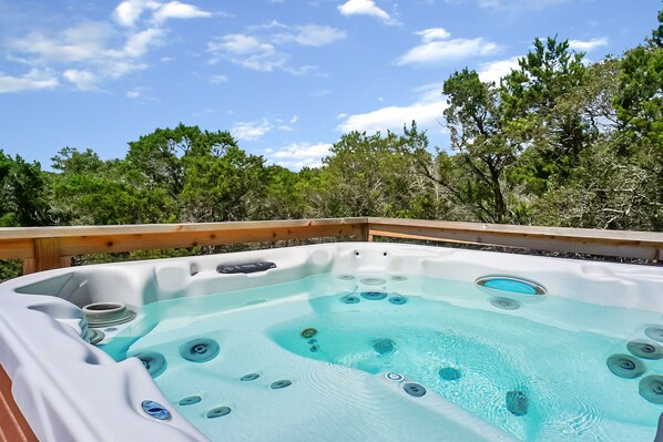 A hot tub with multiple jets, surrounded by a wooden deck and trees, under a clear sky. - Blue Spring Retreat I Hot tub, Families (Wimberley)