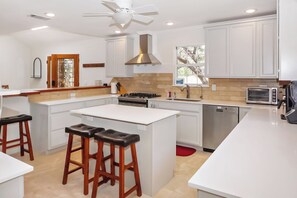 A modern kitchen with white cabinetry, a central island with stools, a built-in stove, a dishwasher, and a ceiling fan.