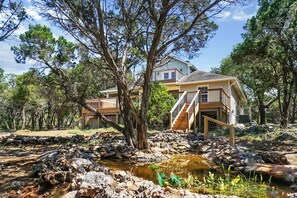A two-story house with a wooden staircase leading to a deck, surrounded by trees and a small stream. - Blue Spring Retreat I Hot tub, Families (Wimberley)