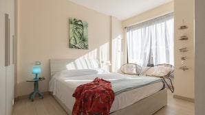 Une chambre à coucher comprenant un lit, une table de chevet avec une lampe, une chaise, une fenêtre avec des rideaux et une œuvre d’art murale.