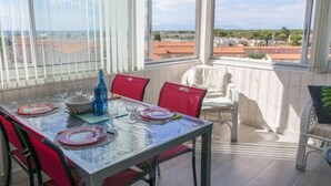 A dining area with a table set for four, red chairs, and a view of the city through large windows. - Apartment Estany by Interhome (Le Barcarès)