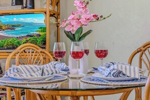 A dining table with a glass top, surrounded by wicker chairs, and a view of a beach through a window. - Classic island condo w/ partial ocean view, & shared hot tub & pool (Kailua-Kona)
