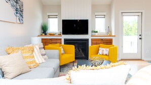 A living room with a grey sofa, yellow armchairs, a flat-screen TV above a fireplace, and a door leading outside. - Discover A Buena Vista on Bald Head Island with stunning views and island vibes. (Bald Head Island)